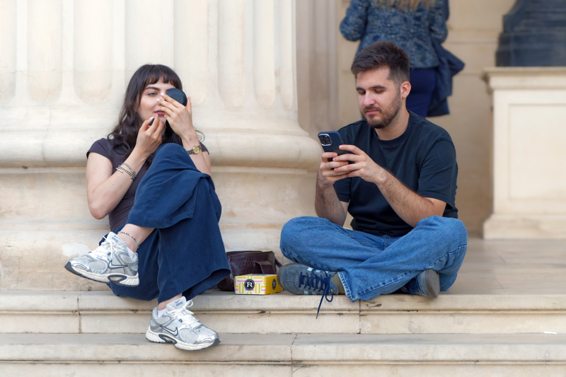 17 - A young woman applying makeup with a compact mirror while wearing dark blue pants and silver sneakers, sitting next to a young man in a dark t-shirt and jeans